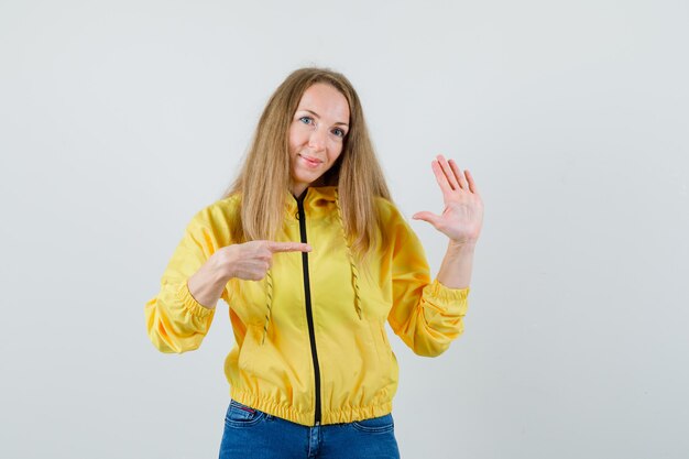 停止ジェスチャーを示し、黄色のボンバージャケットとブルージーンズでそれを指して、楽観的な正面図を探している若い女性。