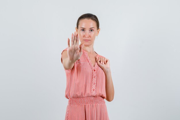 Young woman showing stop gesture in striped dress and looking careful. front view.