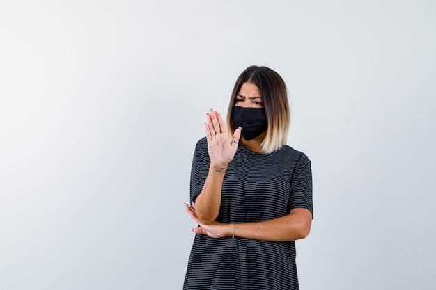 Young woman showing stop sign, holding hand under elbow in black dress, black mask and looking angry. front view.