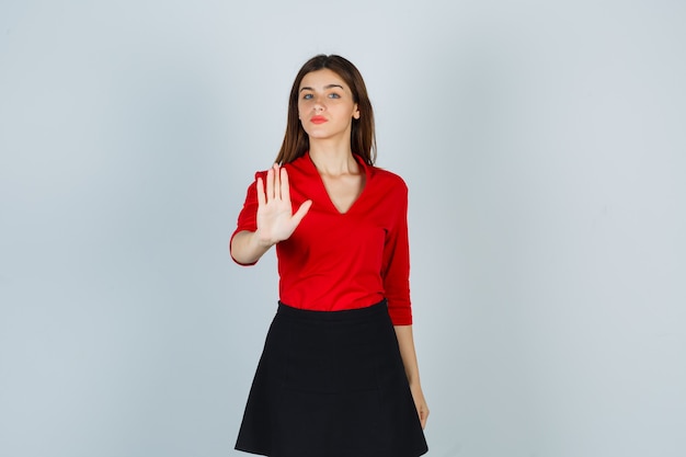 Young woman showing stop sign in red blouse, black skirt and looking serious