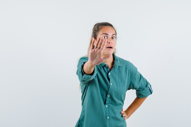 Young woman showing stop sign while holding one hand on waist in green blouse and looking serious