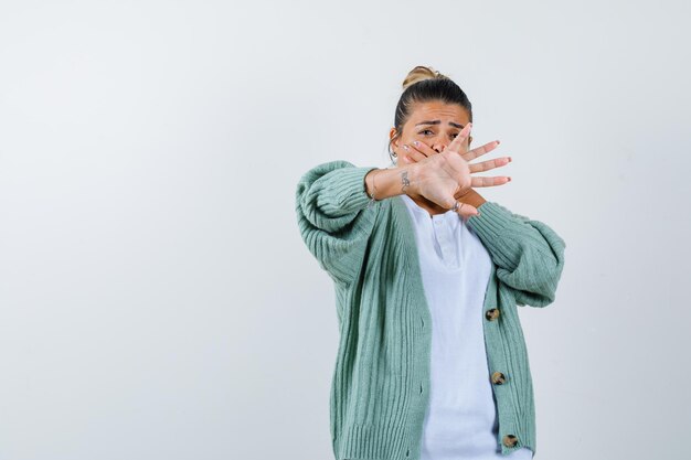 Young woman showing stop sign in white shirt and mint green cardigan and looking scared