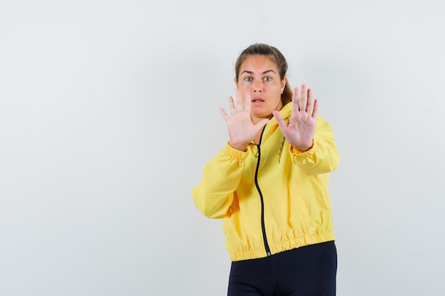 Молодая женщина показывает знак остановки обеими руками в желтой куртке-бомбардировщике и черных штанах и выглядит испуганной