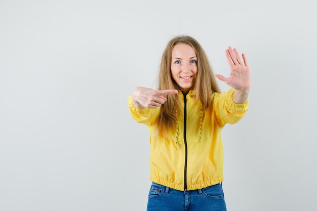 Молодая женщина показывает знак остановки одной рукой и указывает на нее в желтой куртке-бомбардировщике и синих джинсах и выглядит оптимистично, вид спереди.