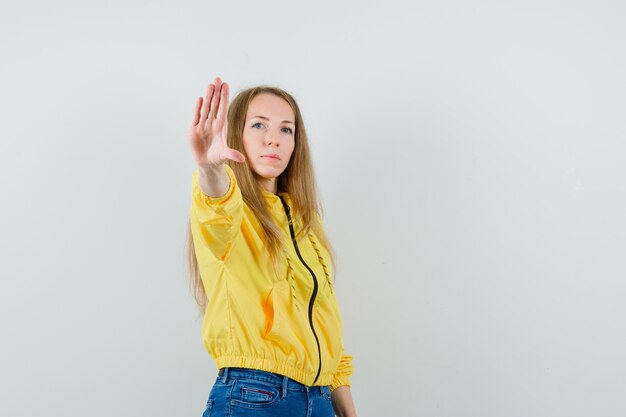 Young woman showing stop sign in yellow bomber jacket and blue jean and looking serious. front view.