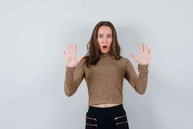 Young woman showing surrender gesture in gold gilded sweater and black pants and looking surprised