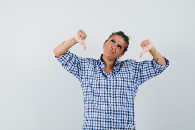 Free photo young woman showing thumb down in checkered shirt and looking displeased.