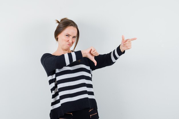 Young woman showing thumb down and pointing right in striped knitwear and black pants and looking displeased