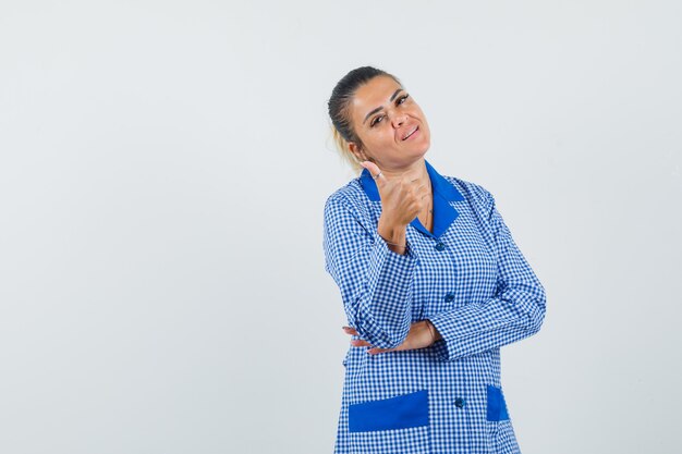 Young woman showing thumb up in blue gingham pajama shirt and looking pretty , front view.