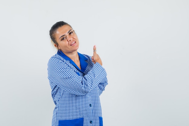 Giovane donna che mostra il pollice in su in camicia pigiama a quadretti blu e sembra carina, vista frontale.