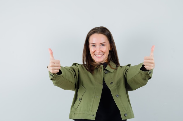 Young woman showing thumb up in green jacket and looking glad. front view.