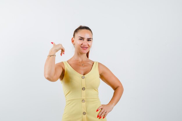 Young woman showing thumb up, holding hand on waist in yellow dress and looking pleased , front view.