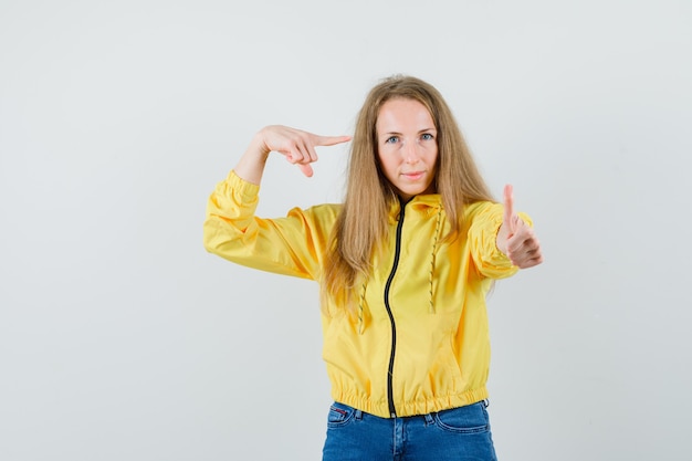 젊은 여자 엄지 손가락을 표시 하 고 노란색 폭격기 재킷과 블루 진에 자신을 가리키고 매력적인, 전면보기를 찾고.
