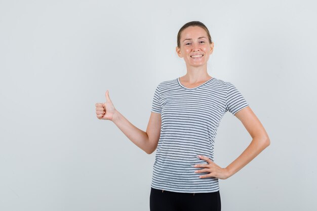Молодая женщина показывает палец вверх в футболке, штанах и выглядит веселым, вид спереди.