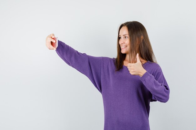 Young woman showing thumb up while taking selfie in violet shirt and looking happy. front view.