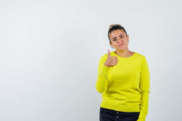 Young woman showing thumb up in yellow sweater and black pants and looking happy