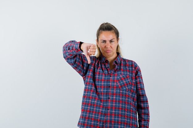 Free photo young woman showing thumbs down in checked shirt and looking displeased