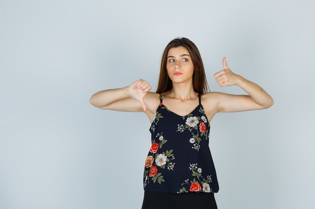 Young woman showing thumbs up and down in floral top and looking uncertain