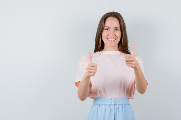 Young woman showing thumbs up in t-shirt, skirt and looking happy. front view.