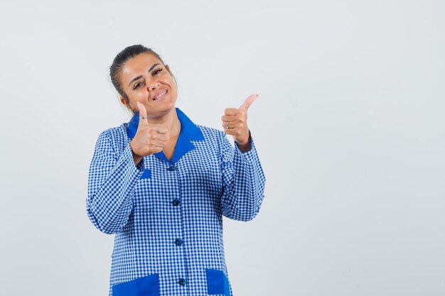 Молодая женщина показывает палец вверх обеими руками в синей пижамной рубашке в клетку и выглядит красиво, вид спереди.