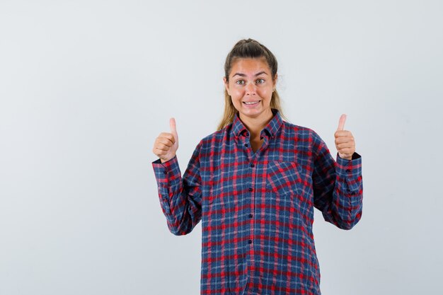 Young woman showing thumbs up with both hands in checked shirt and looking happy