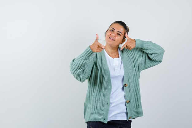 Young woman showing thumbs up with both hands in white t-shirt and mint green cardigan and looking happy