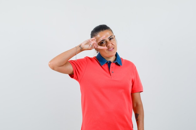 Young woman showing v sign on eye in red t-shirt and looking pretty. front view.