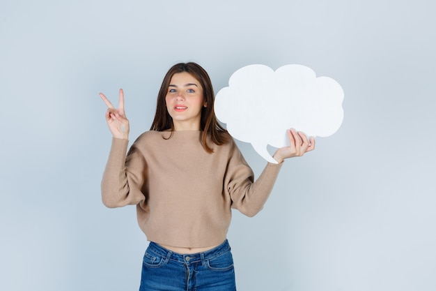 Free photo young woman showing v-sign, keeping paper speech bubble in sweater, jeans and looking wistful , front view.