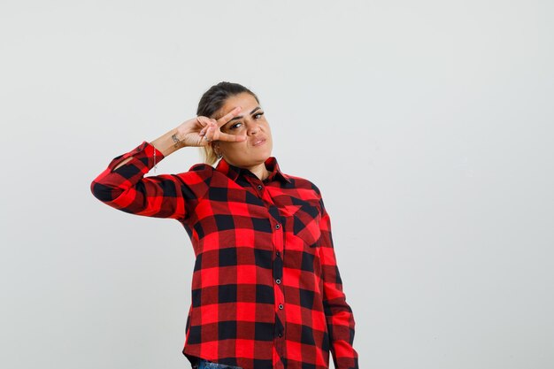 Young woman showing v-sign near eye in checked shirt and looking confident. front view.