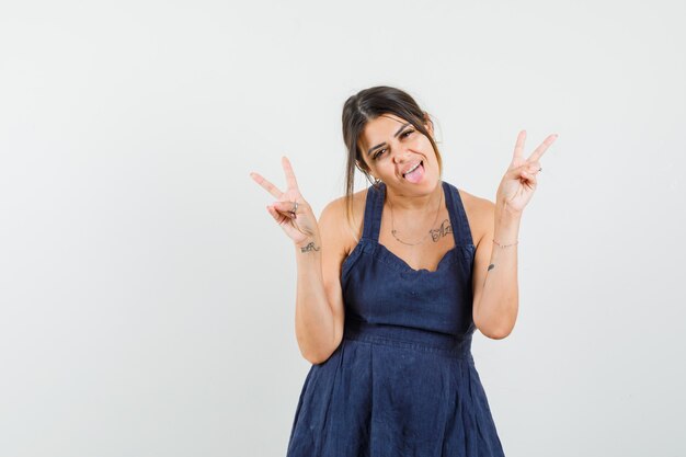 Young woman showing v-sign, sticking out tongue in dress and looking crazy