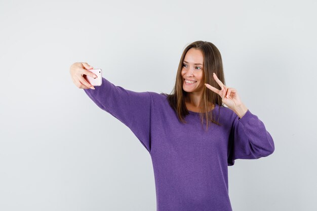 Giovane donna che mostra v-segno mentre prende selfie in camicia viola e sembra felice. vista frontale.