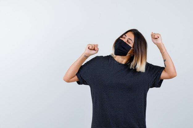 Young woman showing winner gesture in black dress, black mask and looking lucky , front view.