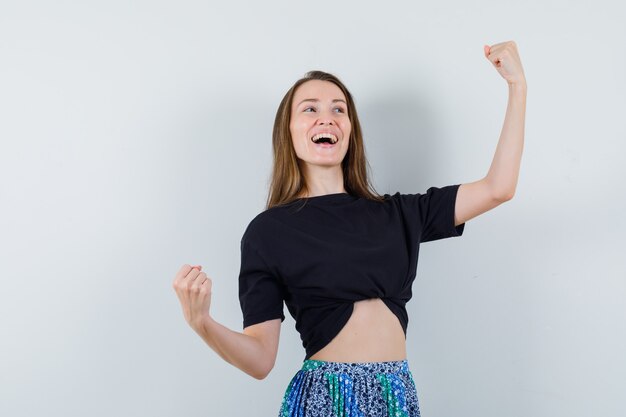 Молодая женщина показывает жест победителя в черной футболке и синей юбке и выглядит счастливой