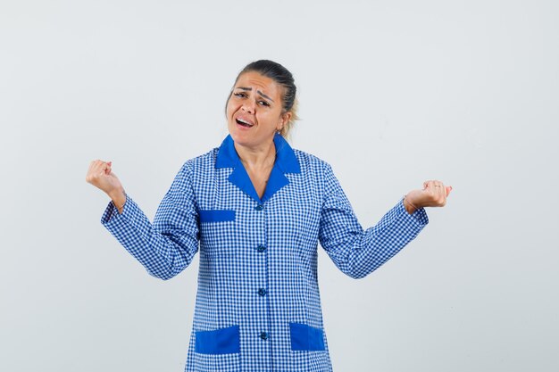 Young woman showing winner gesture in blue gingham pajama shirt and looking excited. front view.