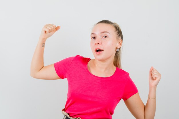 Young woman showing winner gesture in pink t-shirt and looking joyful