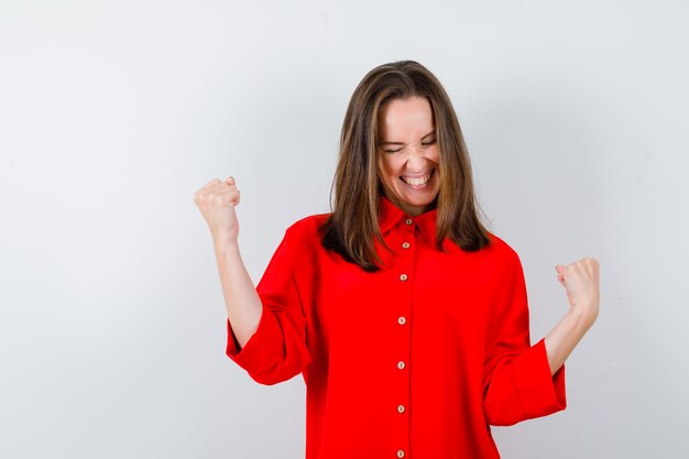 Young woman showing winner gesture in red blouse and looking cheerful , front view.