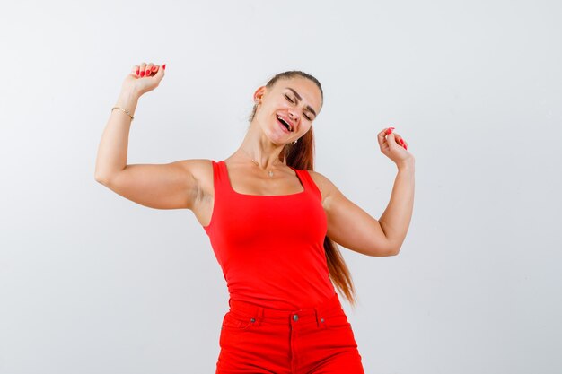 Young woman showing winner gesture in red tank top, pants and looking cheerful , front view.