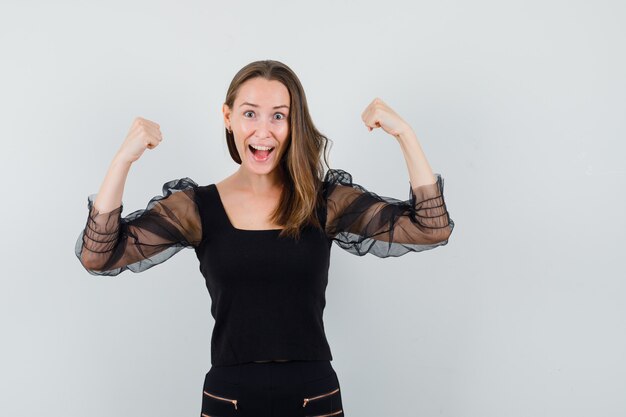 Young woman showing winner pose and smiling joyfully in black blouse and black pants and looking happy 
