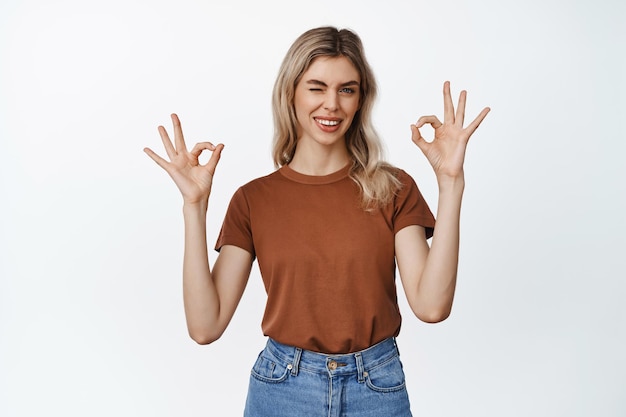 Young woman shows confirmation gesture winking and making okay signs assuring you like and approve product standing over white background