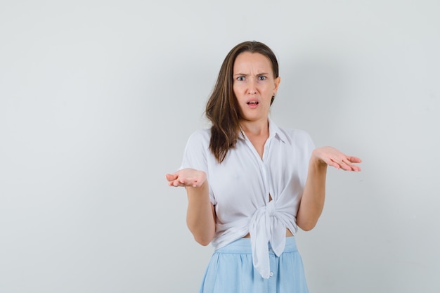 Young woman shrugging shoulders in white blouse and light blue skirt and looking surprised