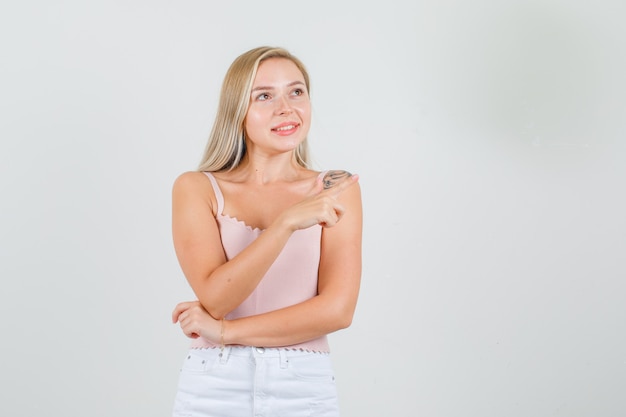 Young woman in singlet, mini skirt looking up while pointing away and looking glad