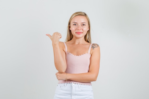 Young woman in singlet, mini skirt pointing to side with thumb and smiling