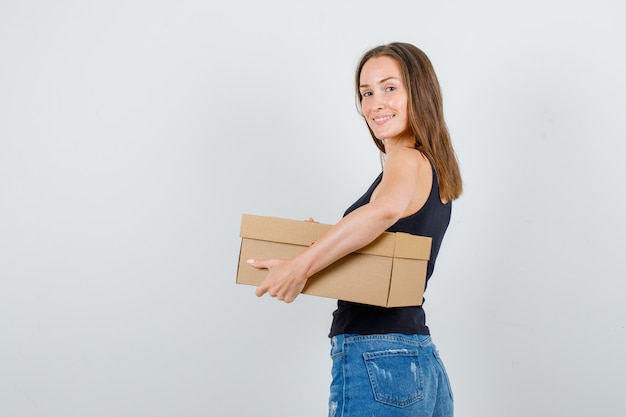 Young woman in singlet, shorts holding cardboard box and looking cheerful .