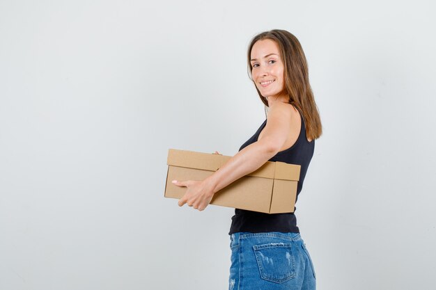 Young woman in singlet, shorts holding cardboard box and looking cheerful .