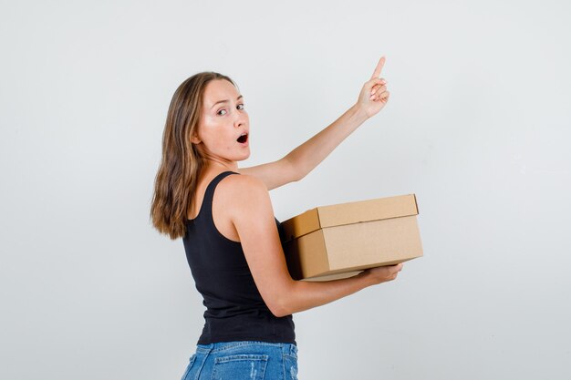 Young woman in singlet, shorts holding cardboard box with finger up .