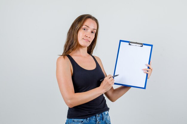 Young woman in singlet, shorts pointing pen at clipboard and looking confident
