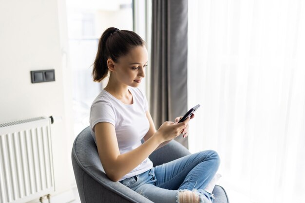 Молодая женщина сидит в кресле и использует смартфон в повседневной одежде в доме в помещении