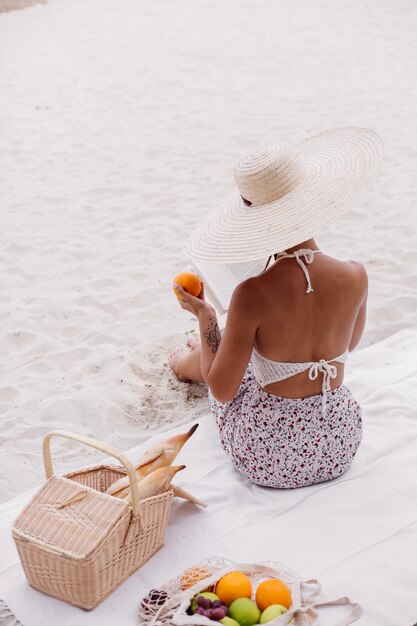 A young woman sits on the beach carpet in a straw hat and a white knitted clothes