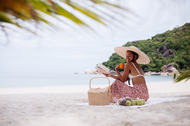 젊은 여자는 밀짚 모자와 흰색 니트 옷에 해변 카펫에 앉아