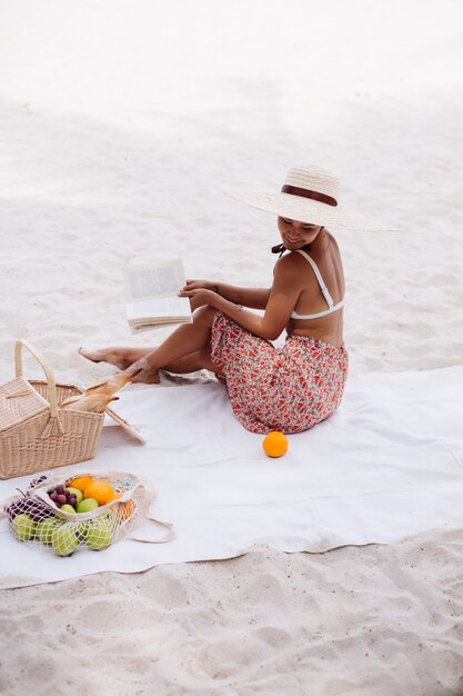 젊은 여자는 밀짚 모자와 흰색 니트 옷에 해변 카펫에 앉아